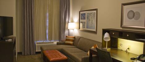 A hotel room with a sofa, ottoman, desk, chair, lamp, and artwork on the walls.
