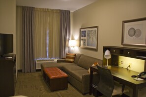 A hotel room with a sofa, ottoman, desk, chair, lamp, and artwork on the walls. - Staybridge Suites Toledo - Rossford - Perrysburg by IHG (Perrysburg)