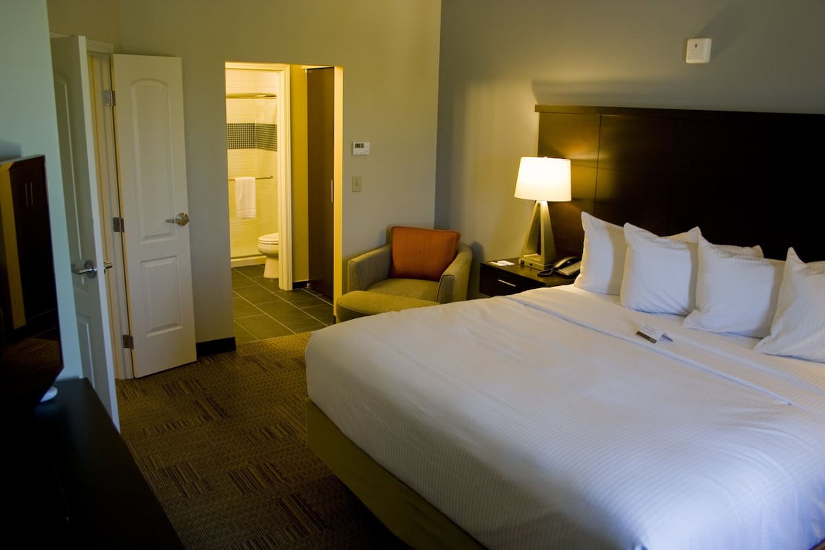 Een hotelkamer met een groot bed, een stoel, een nachtkastje met een lamp, en een badkamer zichtbaar door een geopende deur.