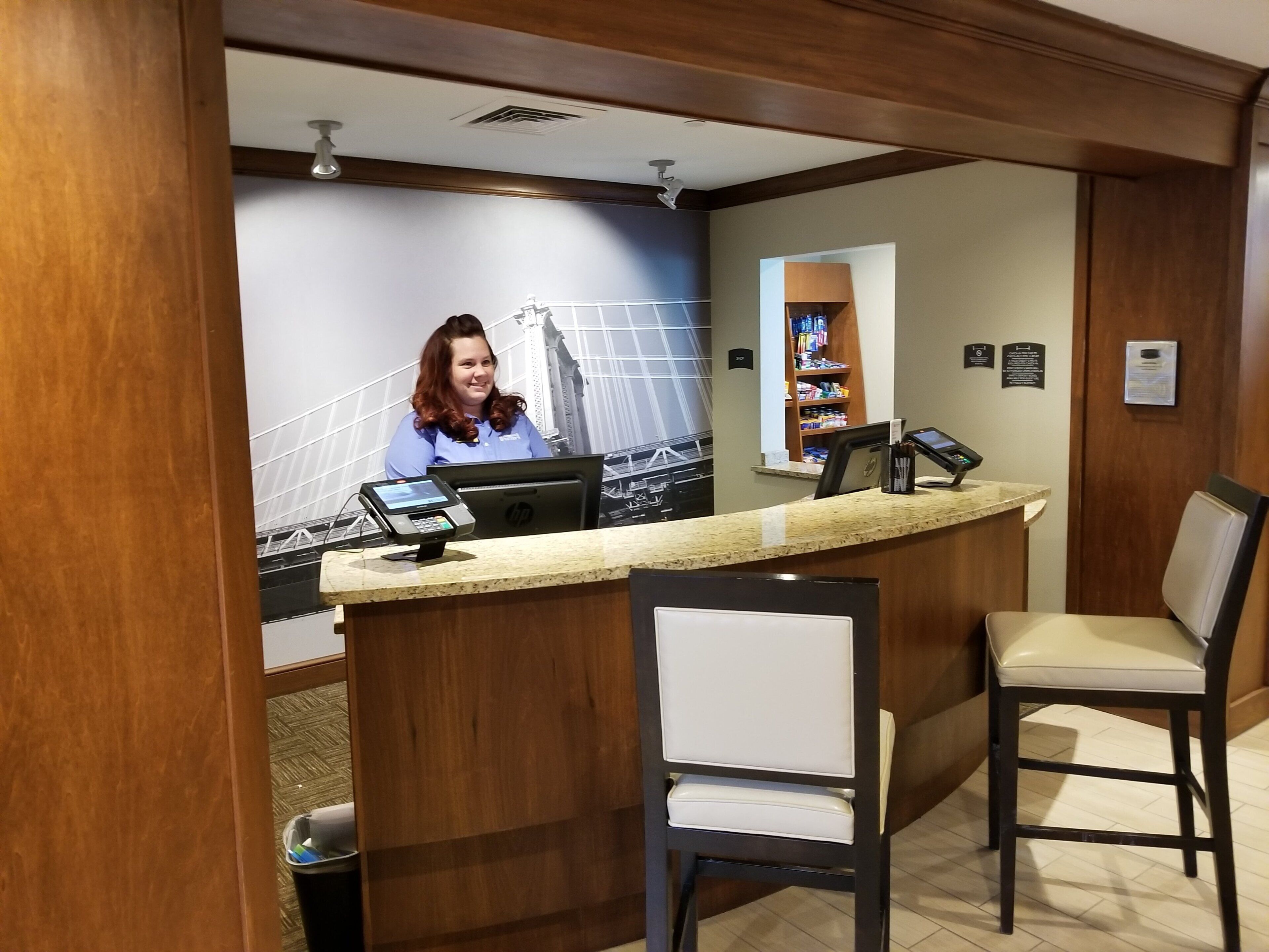 A hotel reception desk with a laptop, a cash register, and a digital screen. There are two chairs in front of the desk, and a wall-mounted shelf with various items.