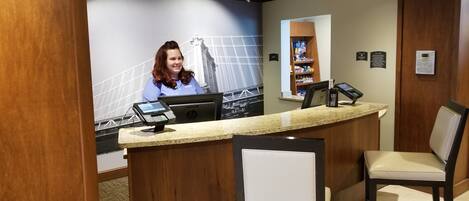 A hotel reception desk with a laptop, a cash register, and a digital screen. There are two chairs in front of the desk, and a wall-mounted shelf with various items.