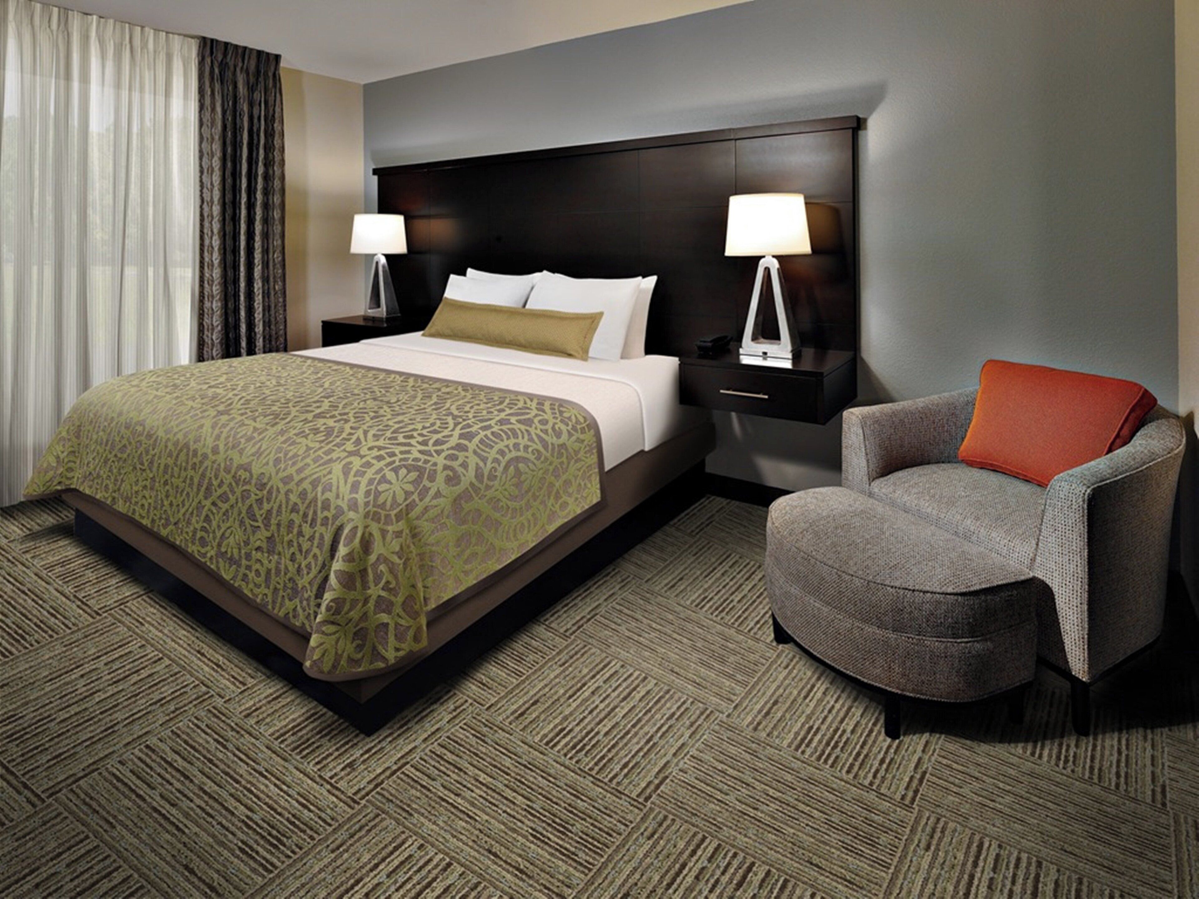 A hotel room with a large bed, two bedside tables with lamps, a grey armchair, and a patterned carpet.