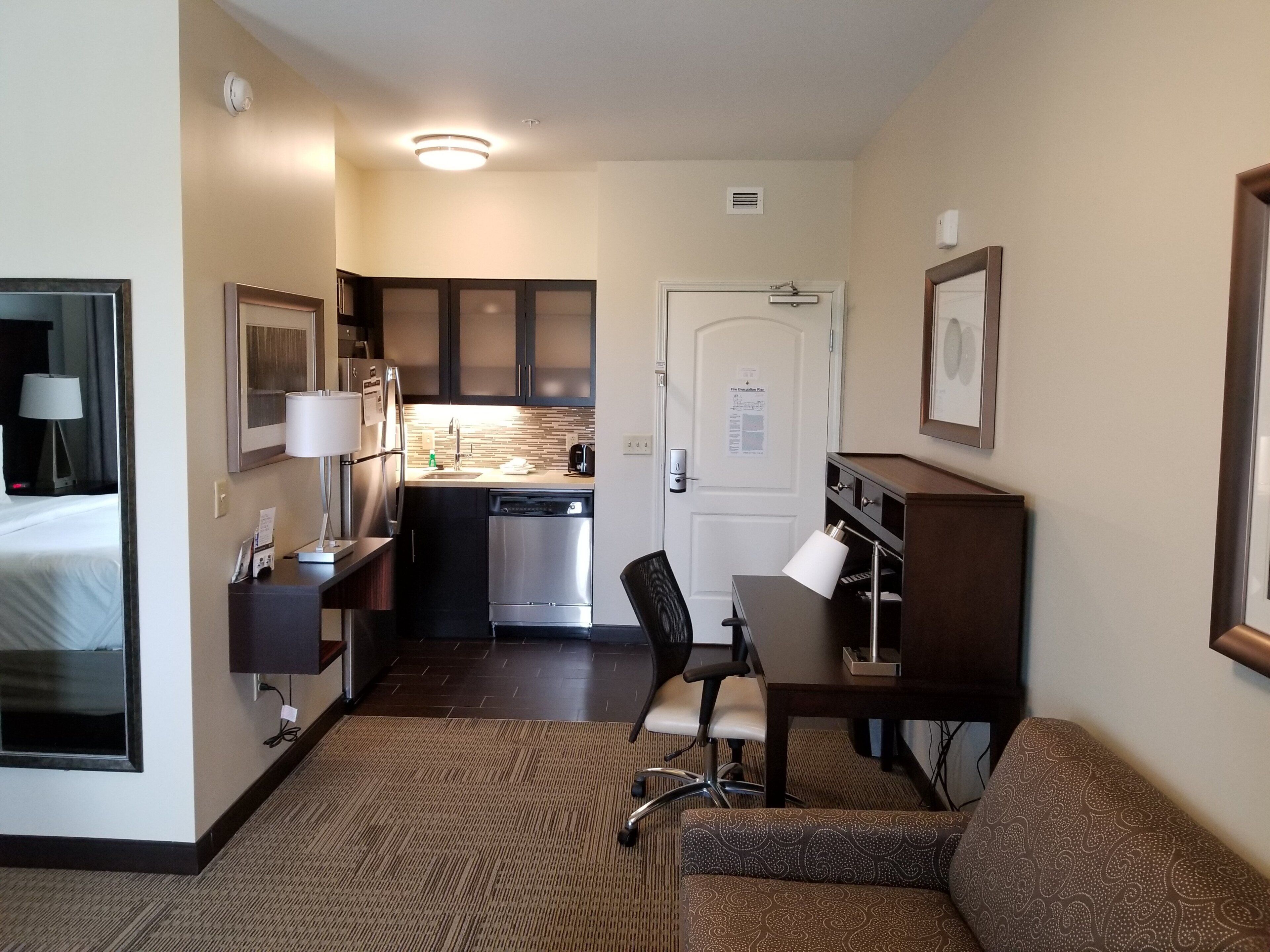Ein kompaktes Hotelzimmer mit Küchenzeile, einem Essbereich mit Tisch und Stühlen, einer Couch, einem Schreibtisch mit Lampe und einem Bett.
