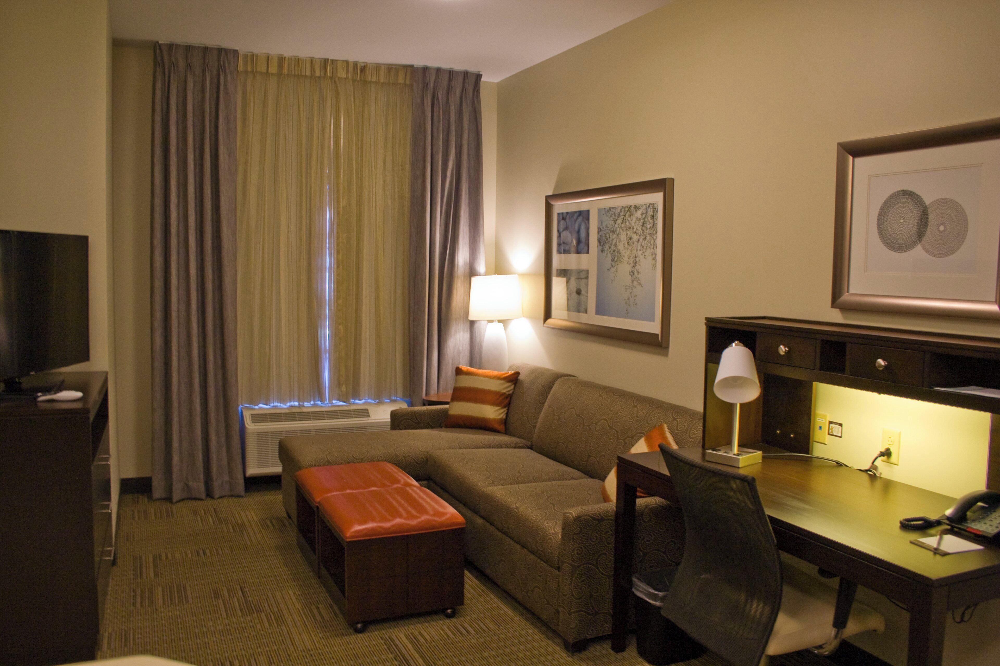 A hotel room with a sofa, an ottoman, a desk with a lamp, a television, and a framed picture on the wall.
