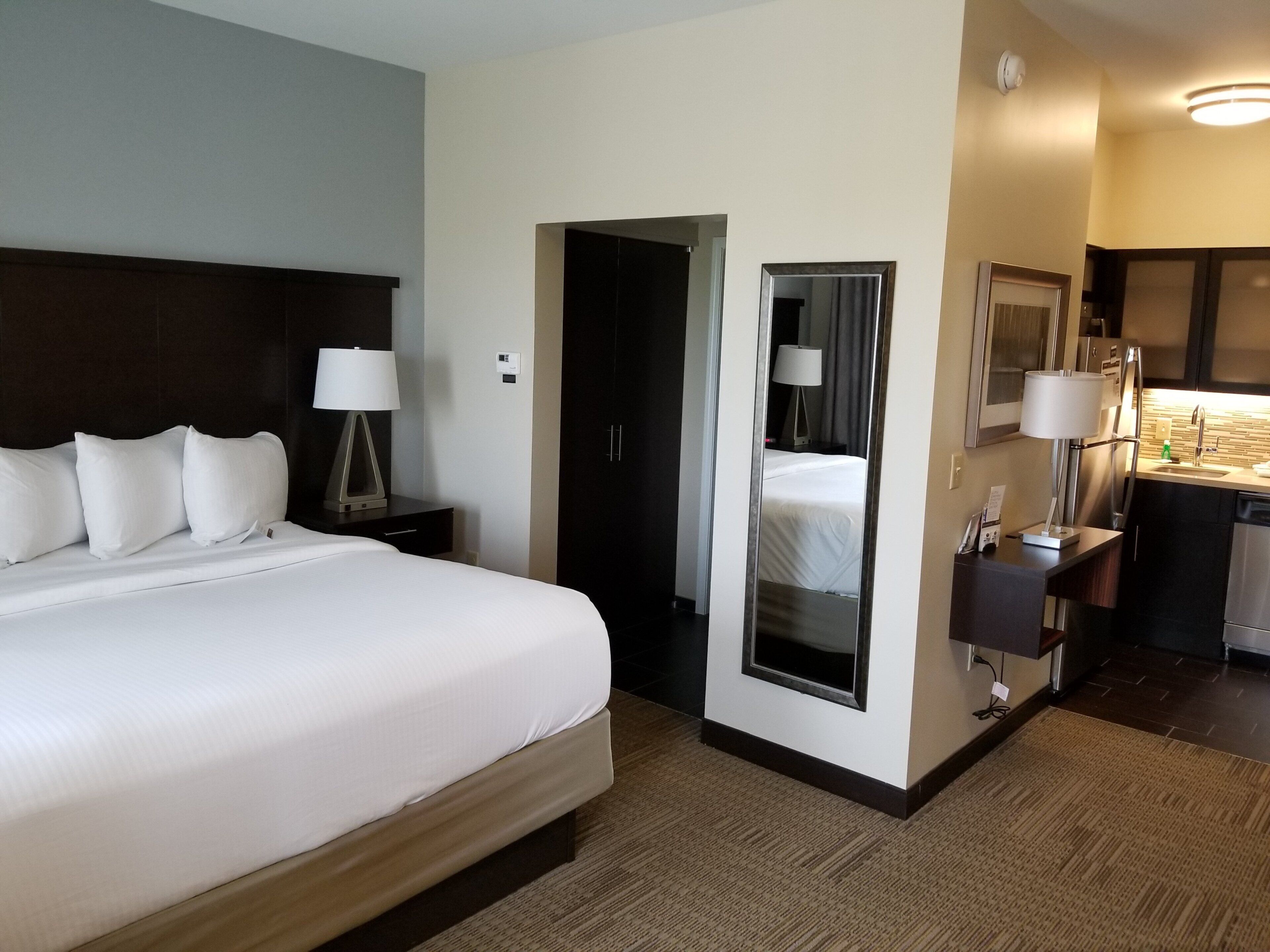 Habitación de hotel con una cama grande, mesitas de noche, un espejo, un escritorio con lámpara y una kitchenette.