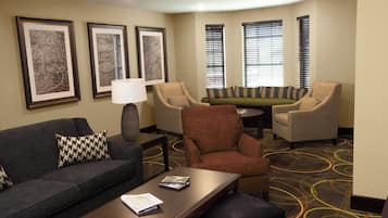 Ein Hotelzimmer mit einer Couch, Sesseln, einem Couchtisch und einer Lampe. An den Wänden hängen Bilderrahmen und der Boden ist mit Teppich ausgelegt.
