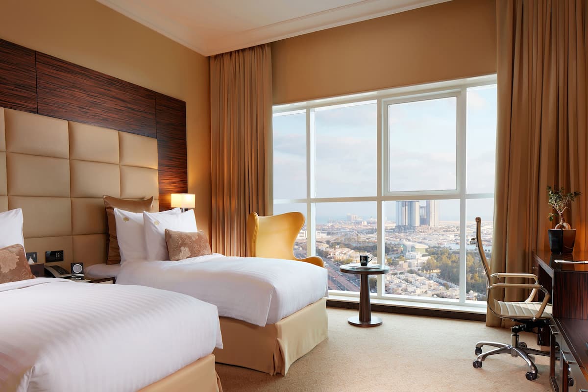 A hotel room with two beds, a desk, a chair, a view of the city, and a large window.