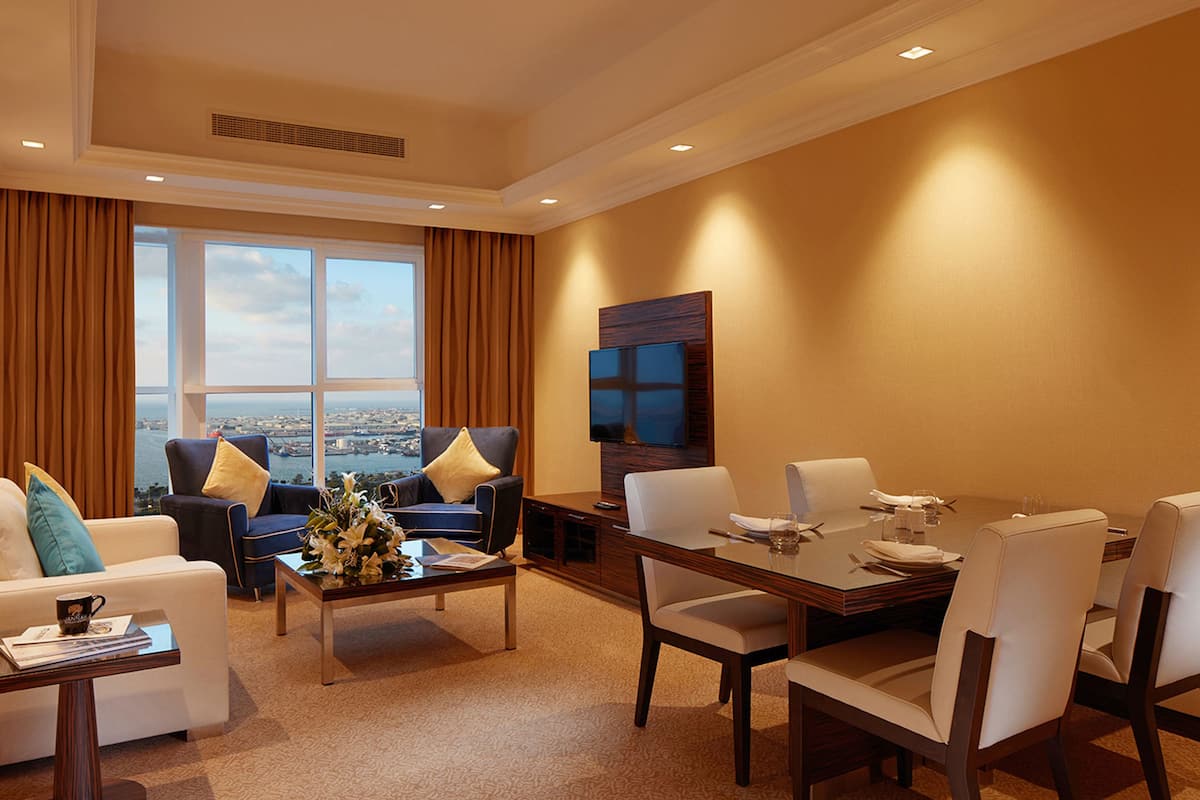 A hotel room with a large window offering a city view, a dining area with a table set for four, and a living area with a sofa and armchairs.