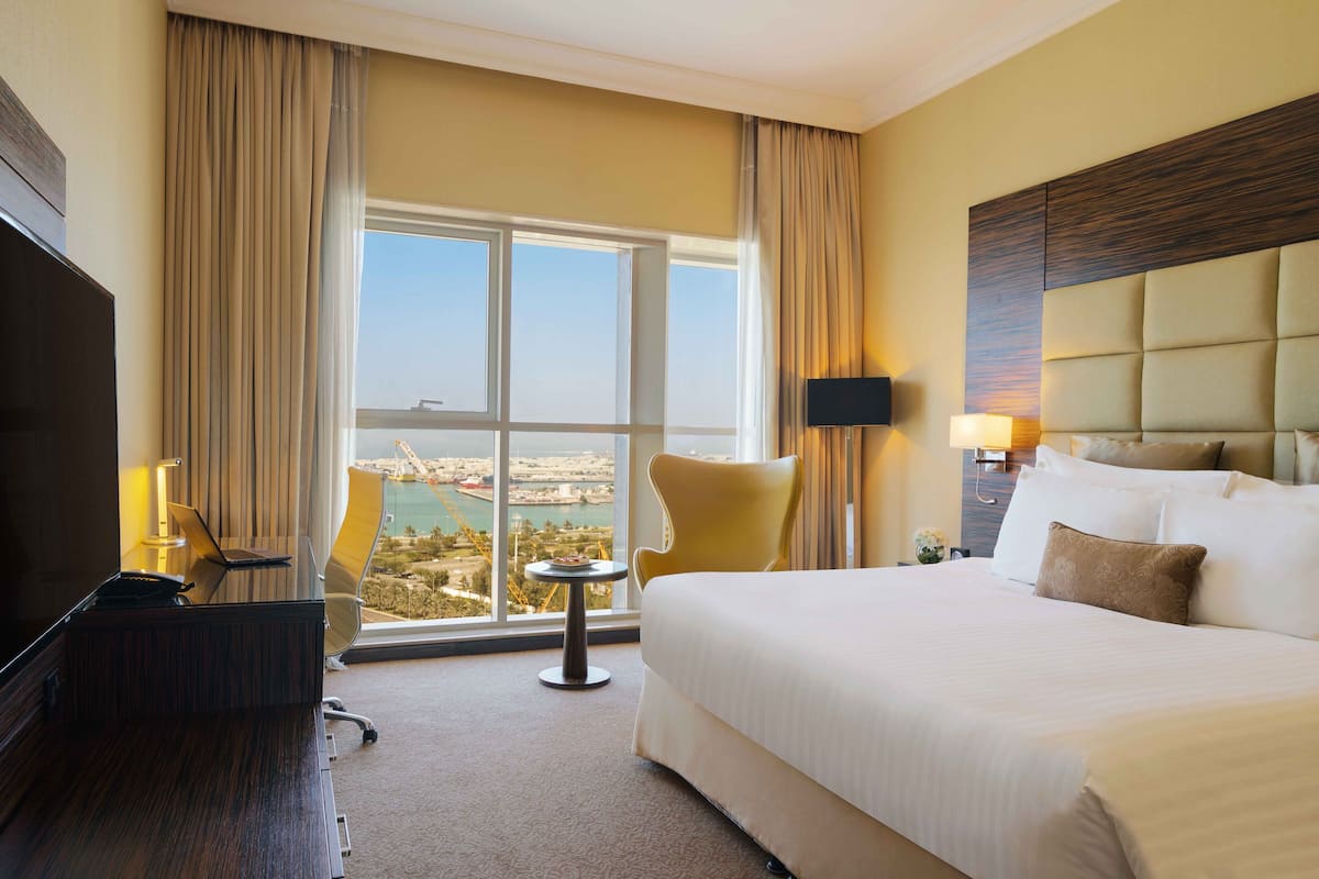 A hotel room with a large bed, a desk with a laptop, a chair, and a view of a cityscape through a large window.