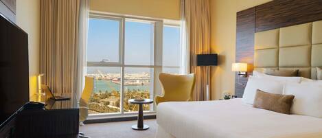 A hotel room with a large bed, a desk with a laptop, a chair, and a view of a cityscape through a large window.