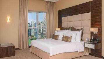 A hotel room with a large bed, a nightstand, a lamp, and a view of tall buildings through a window.