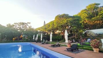 A swimming pool with lounge chairs and umbrellas, surrounded by greenery and a view of a hill.
