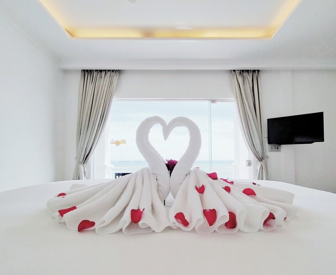 Une chambre d’hôtel avec un lit, une décoration en forme de cœur faite de serviettes et de pétales de rose, et un téléviseur fixé au mur.