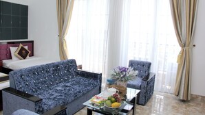 Habitación de hotel con cama, dos sofás, una mesa de centro con fruta y un jarrón, y cortinas translúcidas.