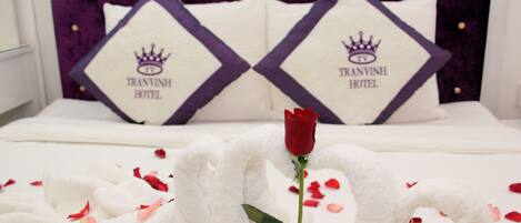 A bed with white bedding, two pillows with "TRAVINH HOTEL" and a crown logo, and a single red rose.