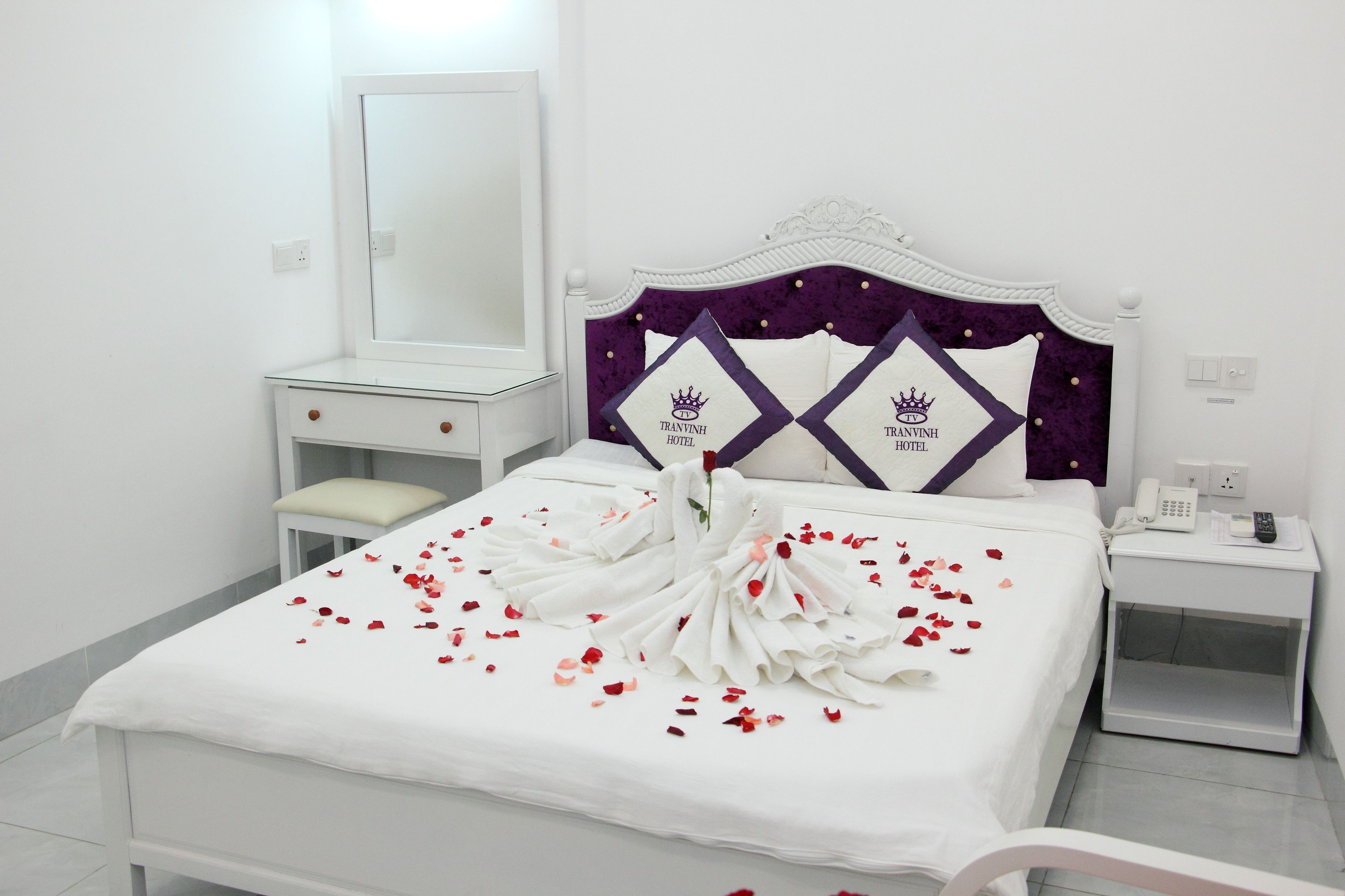Una cama bien hecha con una colcha blanca y un cabecero morado, una mesita de noche con un teléfono y un espejo en la pared.