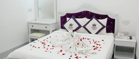 Una cama bien hecha con una colcha blanca y un cabecero morado, una mesita de noche con un teléfono y un espejo en la pared.