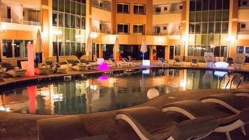 An indoor swimming pool with lounge chairs and umbrellas, surrounded by a multi-story building with large windows.