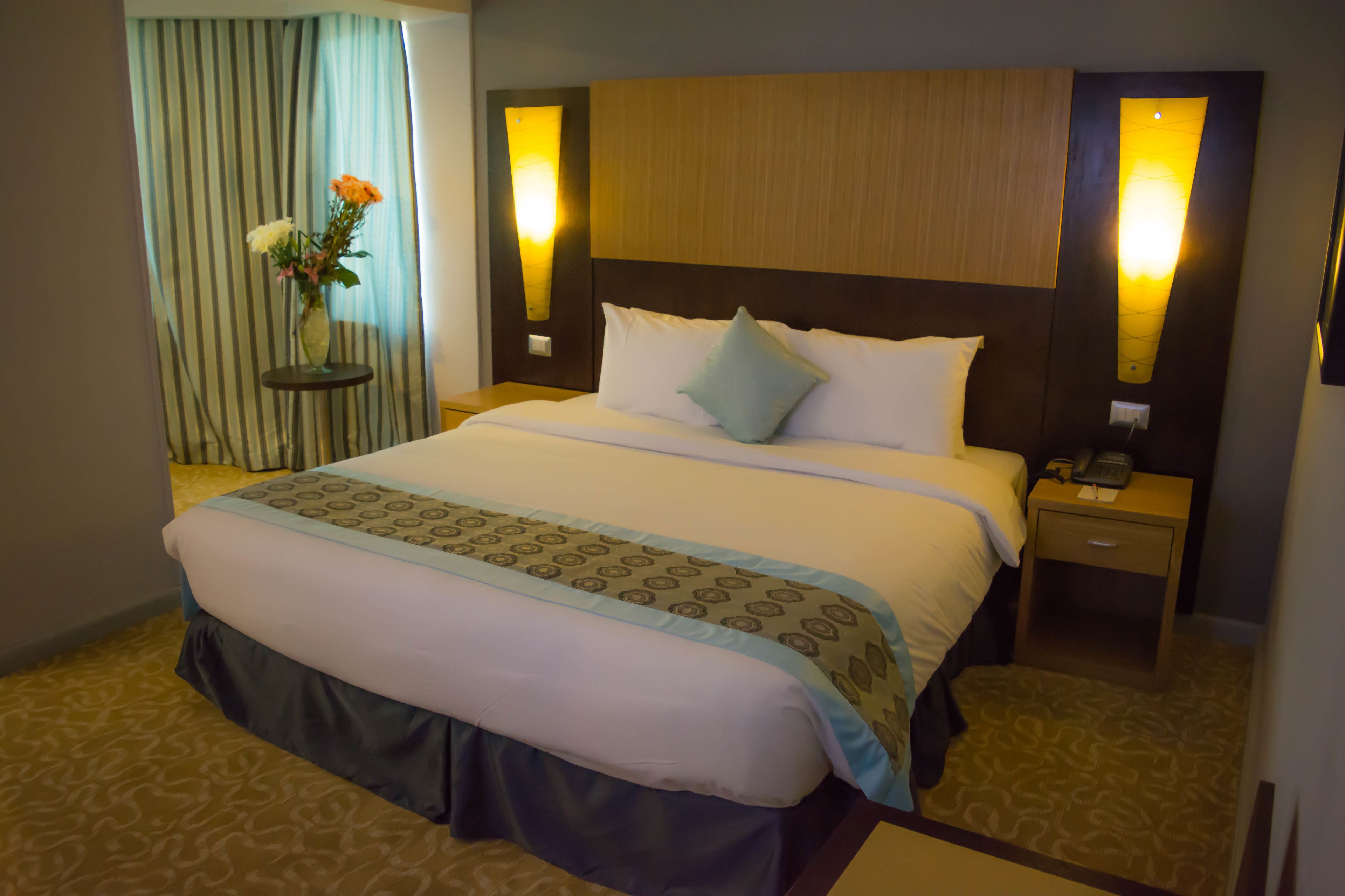Une chambre d’hôtel avec un grand lit, une table de chevet, un vase de fleurs et un rideau.