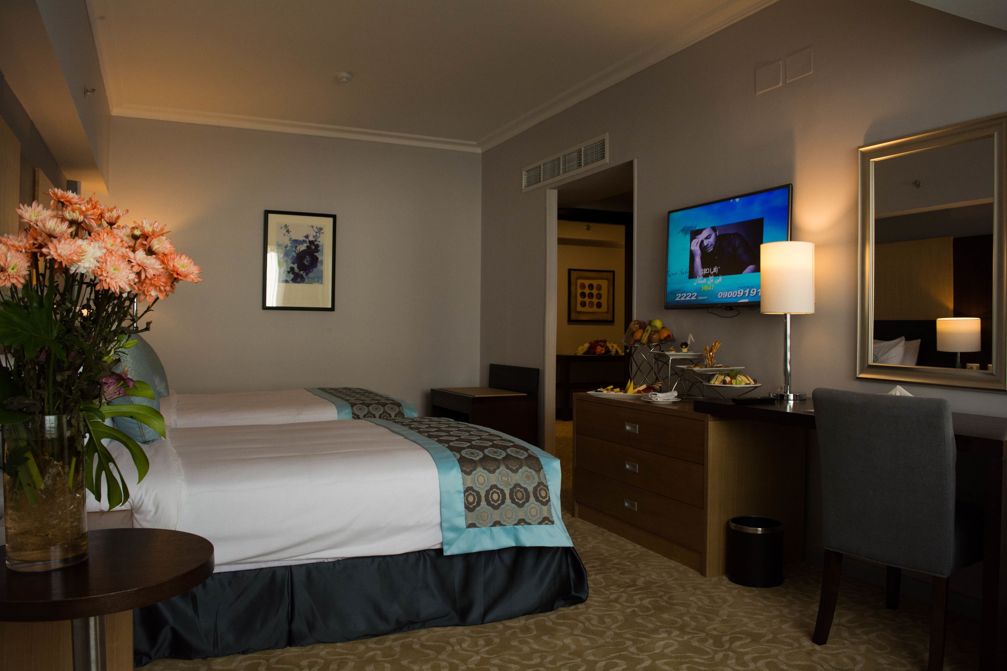A hotel room with a bed, a desk, a chair, a TV, a lamp, and a vase of flowers.