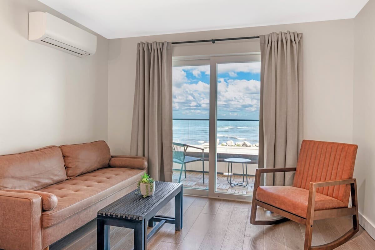 A room with a sofa, a chair, a small table, and a view of the ocean through large windows.