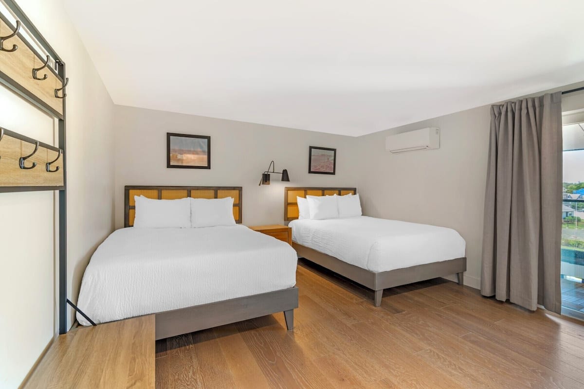 A hotel room with two beds, a wooden floor, a balcony with curtains, and a wall-mounted air conditioning unit.