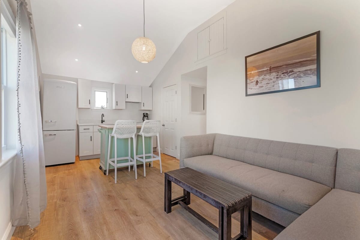 A modern living room with a grey sofa, a wooden coffee table, and a kitchen area with white cabinets and a bar stool.