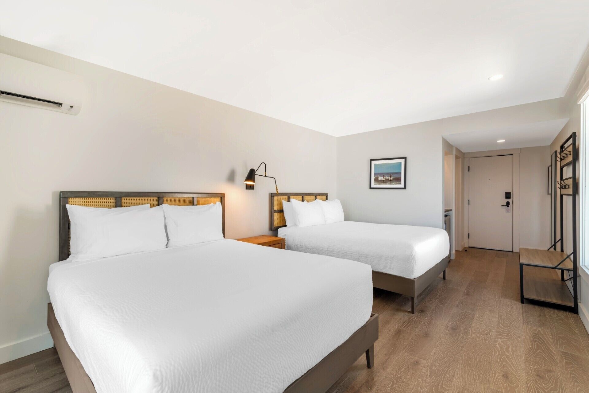 A hotel room with two beds, a bedside table with a lamp, a picture on the wall, and a door leading to another room.