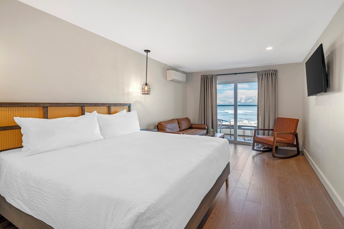 A hotel room with a large bed, a seating area with a view of the sea, and a flat-screen TV mounted on the wall.