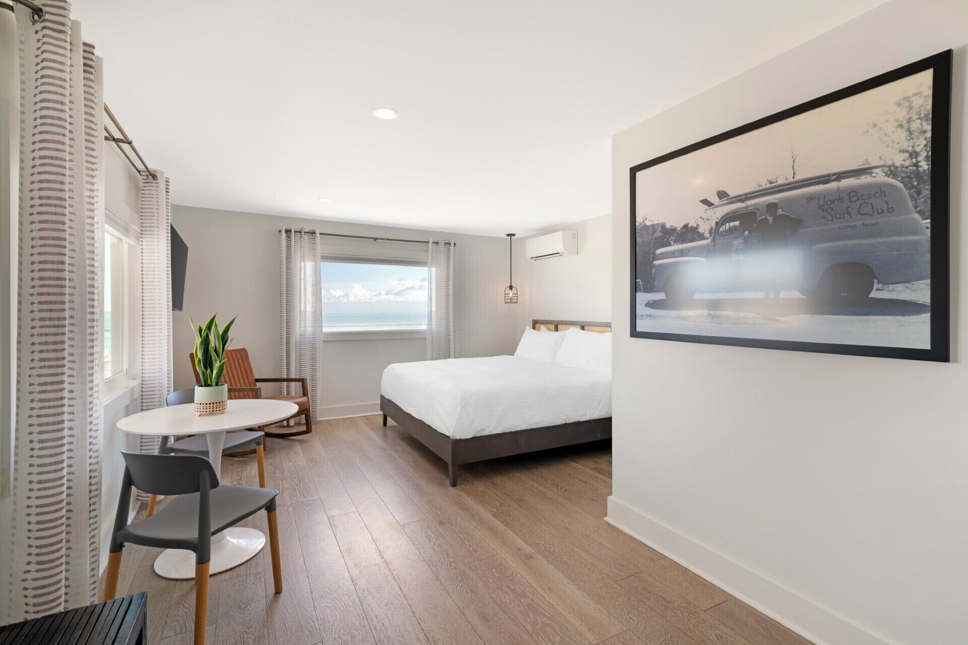 A hotel room with a bed, a desk, a chair, a potted plant, a window with curtains, and a framed picture on the wall.