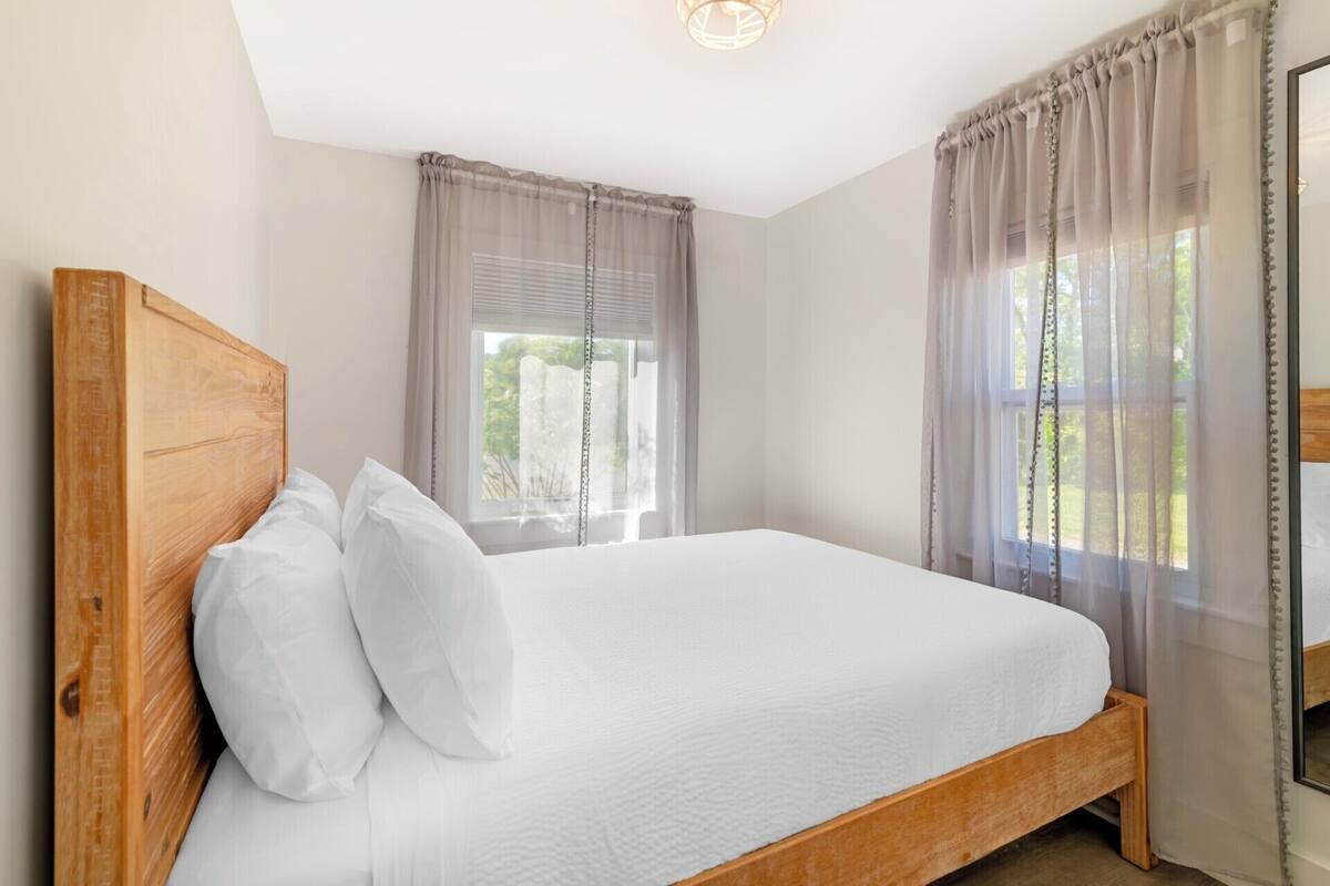 A bedroom with a wooden headboard, a white bedspread, and sheer curtains on the windows.