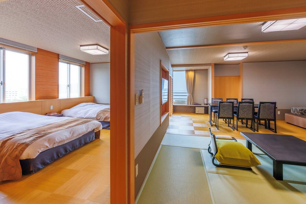 A hotel room with two beds, a dining area with a table and chairs, and a view of the sea through the window.