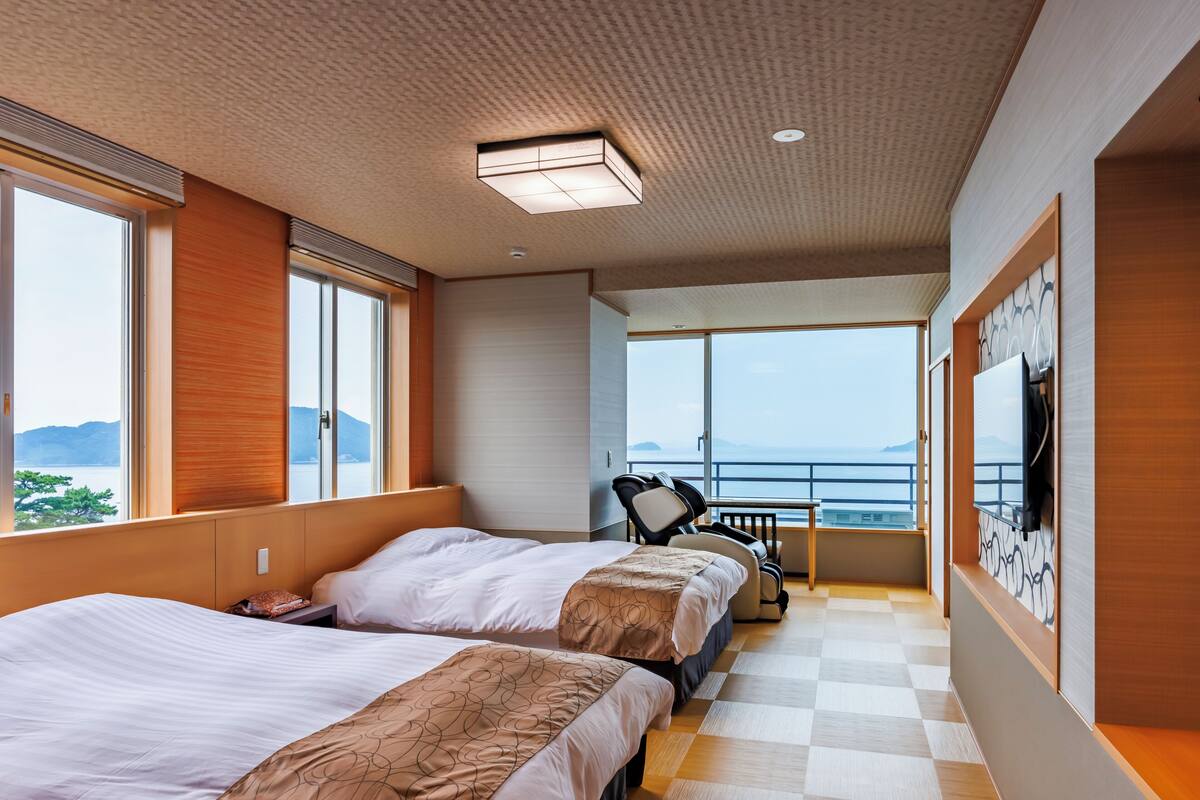 A hotel room with two beds, a massage chair, a balcony with a view of the sea, and a large window.