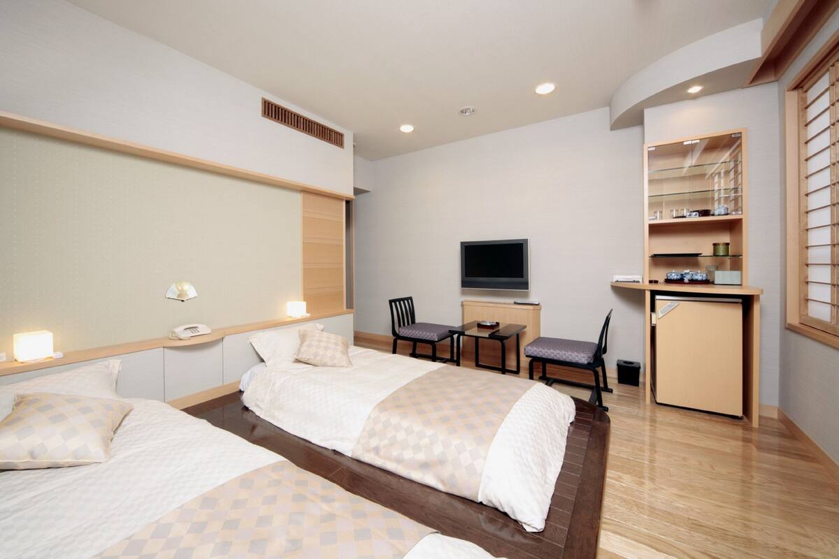 A hotel room with two beds, a desk, a TV, and a kitchenette.