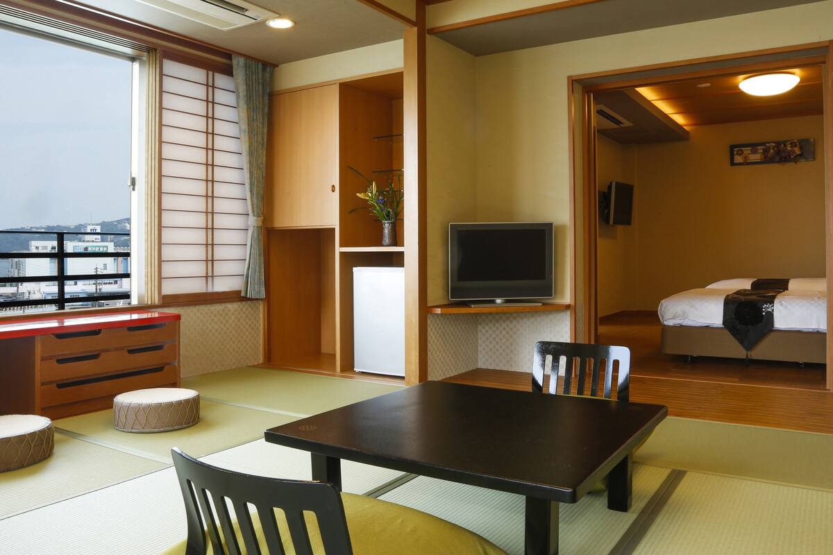 A traditional Japanese room with tatami flooring, a wooden table, chairs, a television, and a bed.