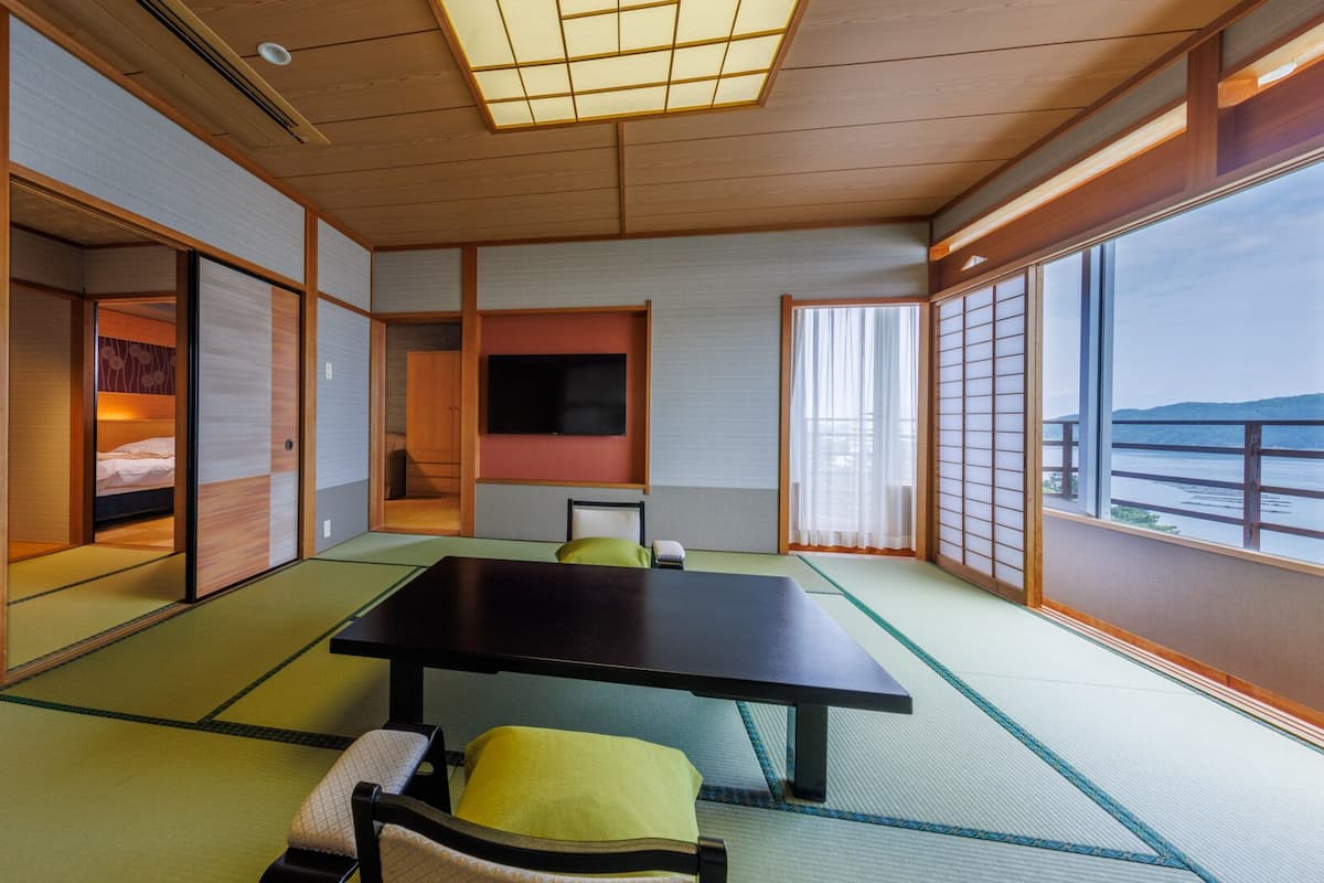 A traditional Japanese room with tatami flooring, a low table, a bed with a futon, sliding doors, and a view of the water.