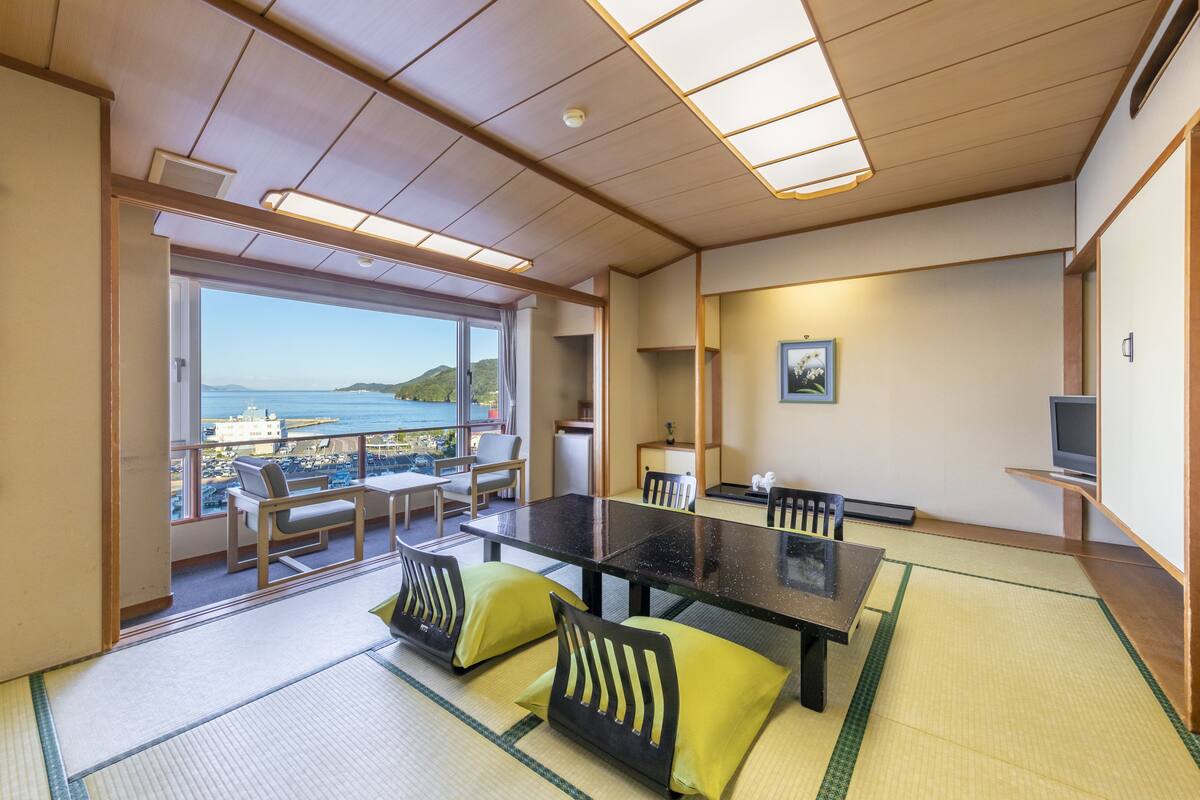 A room with a large table, chairs, tatami mats, a TV, and a view of the sea.