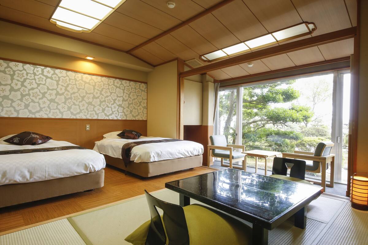 A hotel room with two beds, a dining table, chairs, and a view of greenery outside.