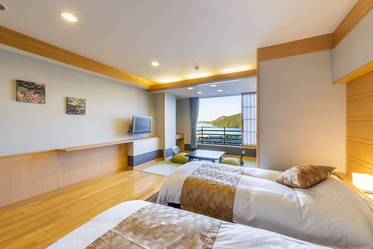 A hotel room with two beds, a TV, a balcony with a view of hills, and a wooden floor.