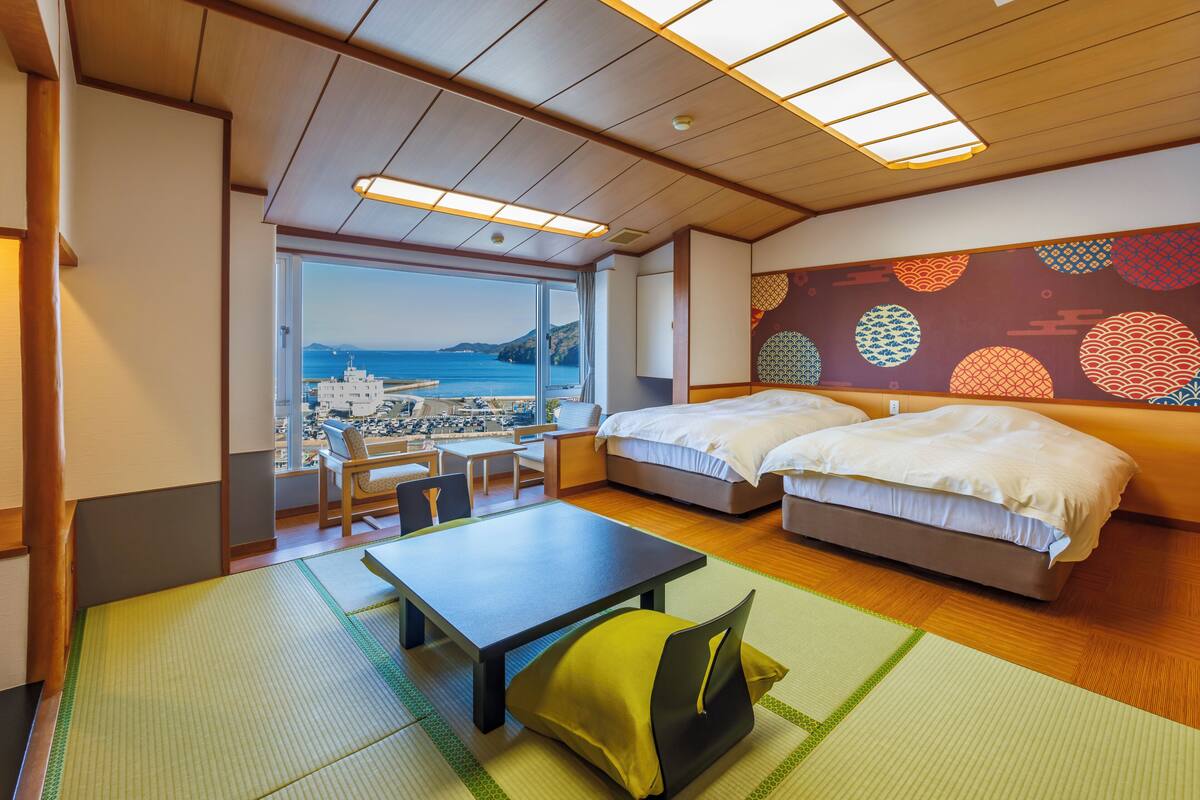 A hotel room with a large window, two beds, a table, and a view of the sea.