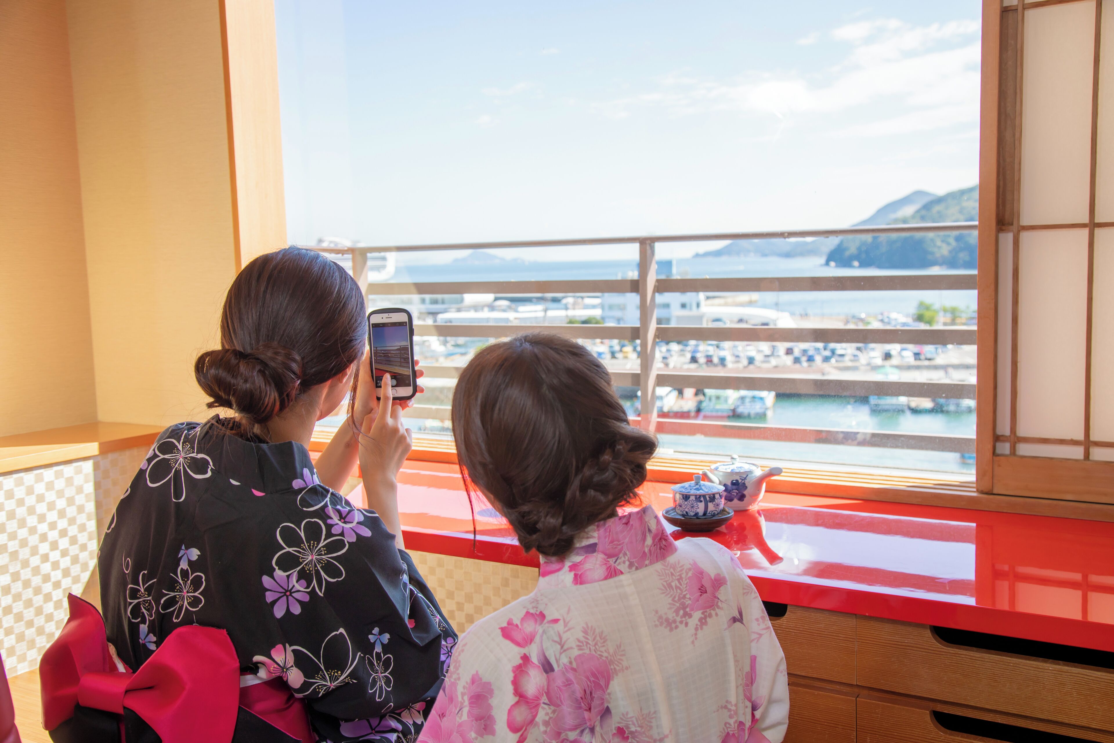 Two people taking a photo of a coastal view from a balcony with a red counter and a traditional Japanese sliding door.