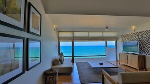 A modern living room with a large window overlooking the sea, framed artwork, a television, and a wooden cabinet. - Pandanus Beach Resort & Spa (Induruwa)