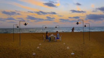A beachside dining setup with a table and chairs, surrounded by sand and ocean views.