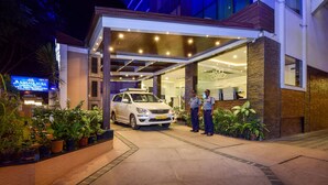 A modern building with a covered entrance, a white car parked in front, and security personnel standing nearby. - Akshaya Aura (Bengaluru)