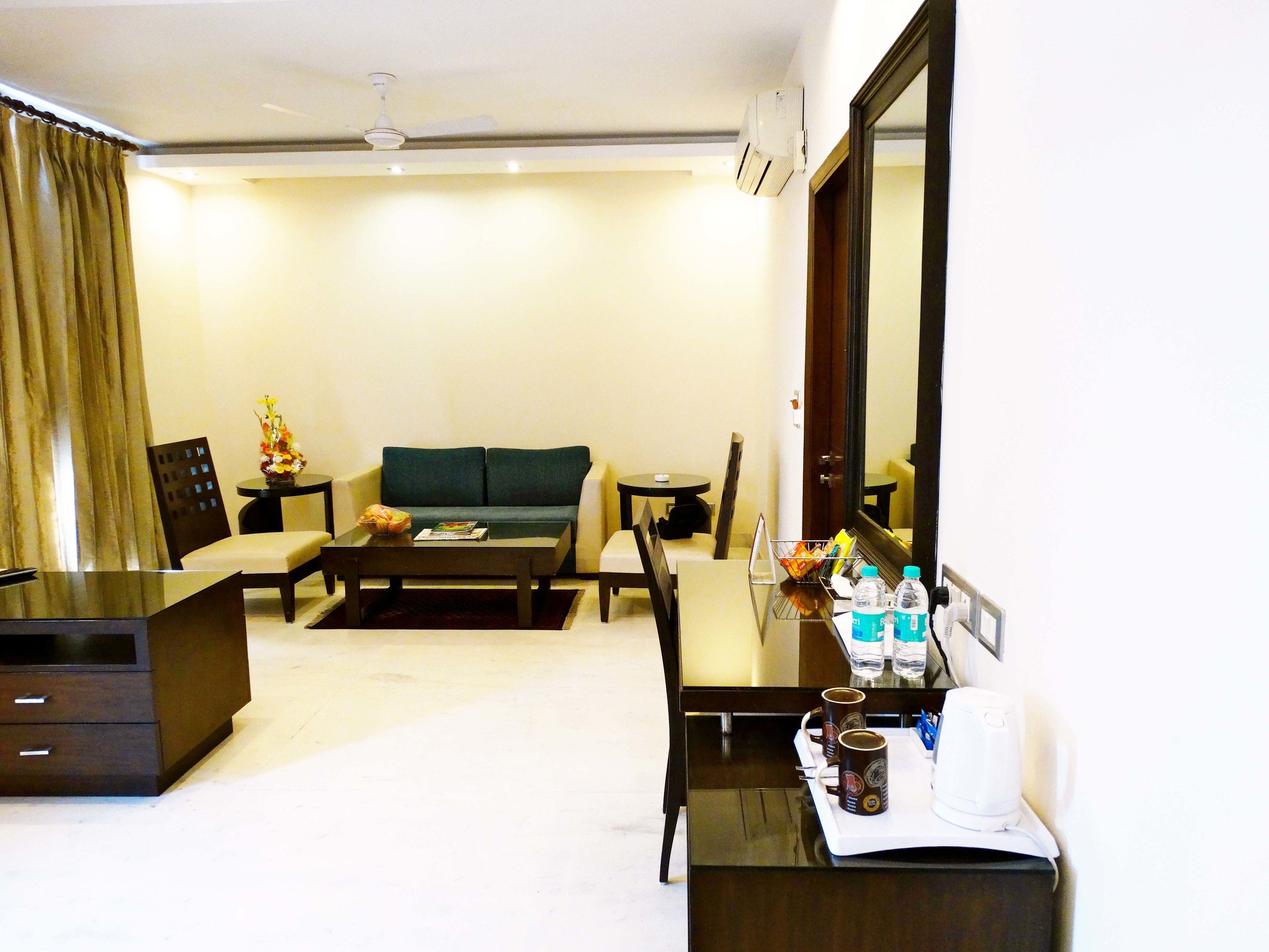 A hotel room with a sofa, a coffee table, a TV stand, and a desk with a coffee maker and cups.