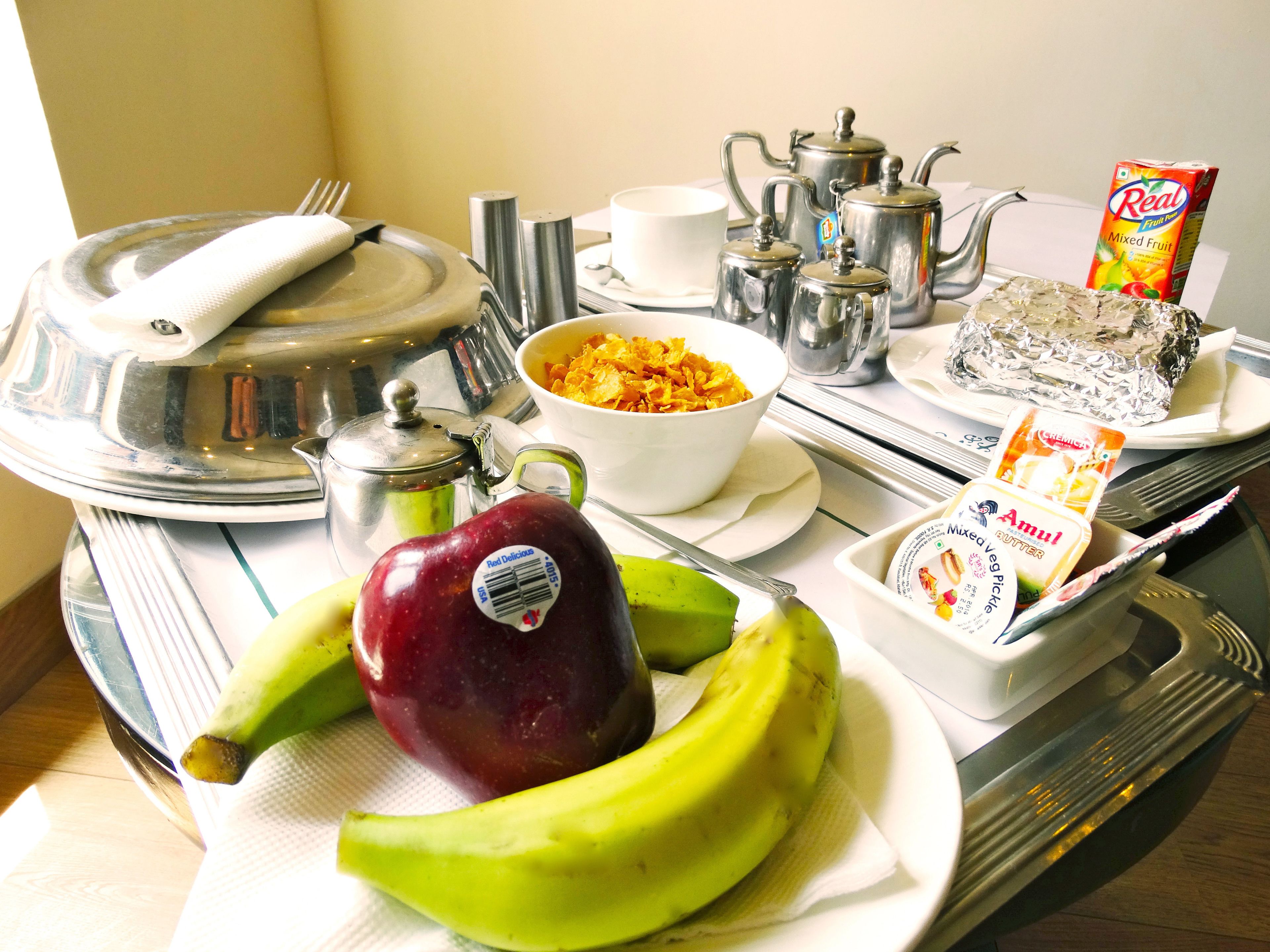 A breakfast tray with a red apple, bananas, a bowl of cereal, a carton of mixed fruit juice, a container of Amul butter, and a teapot.