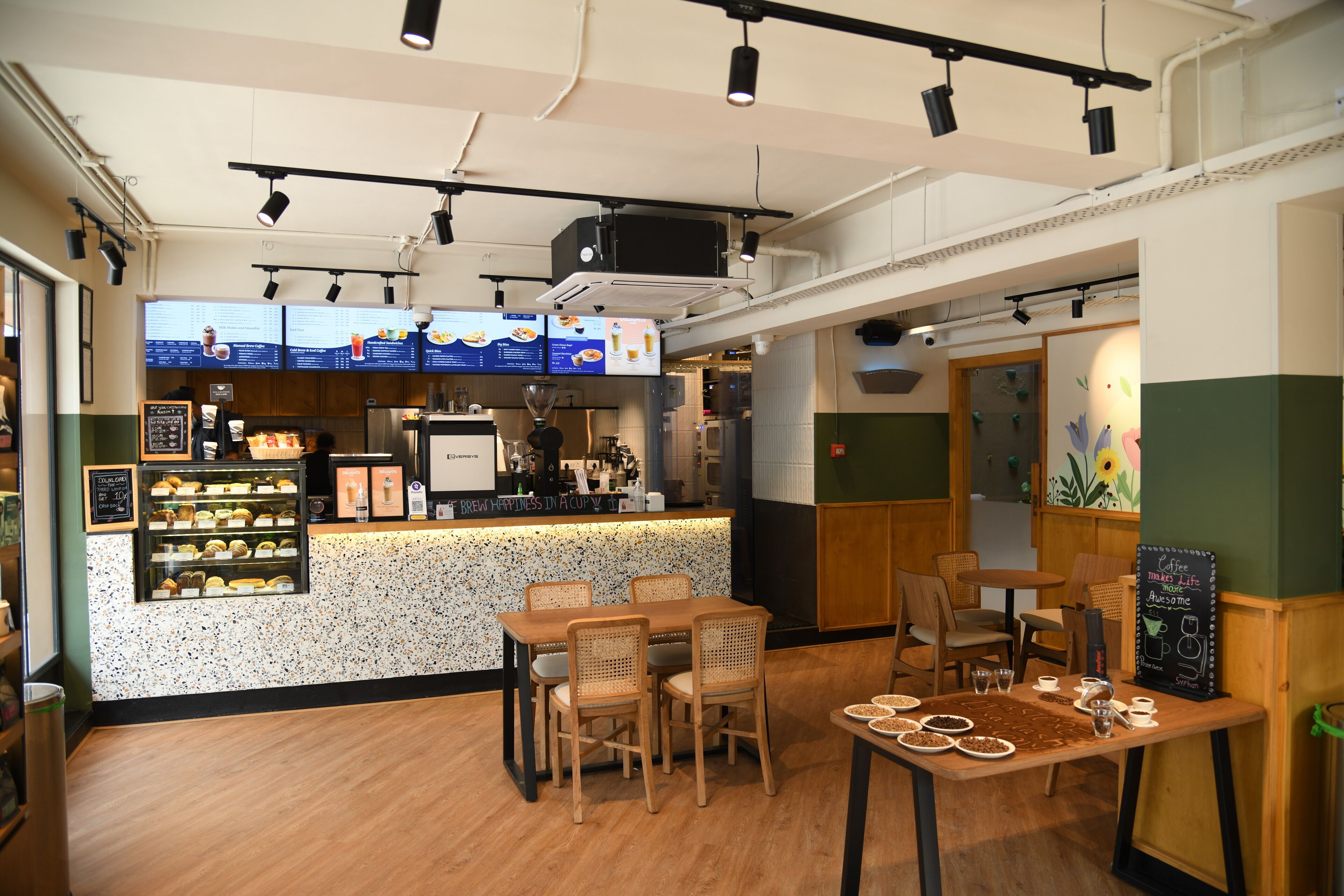 A café with a counter displaying menu items, wooden tables and chairs, and a chalkboard sign.
