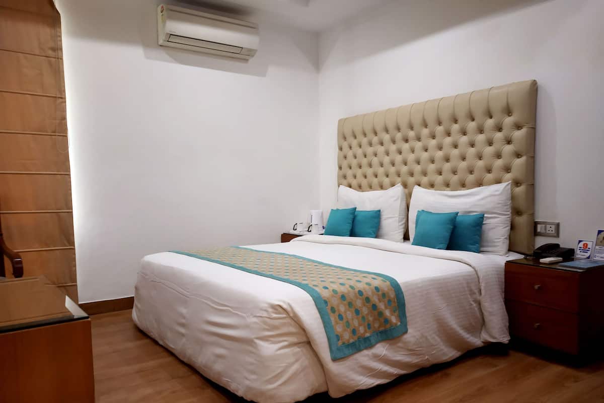 A hotel room with a large bed, a wooden nightstand, a beige upholstered headboard, and a wall-mounted air conditioner.