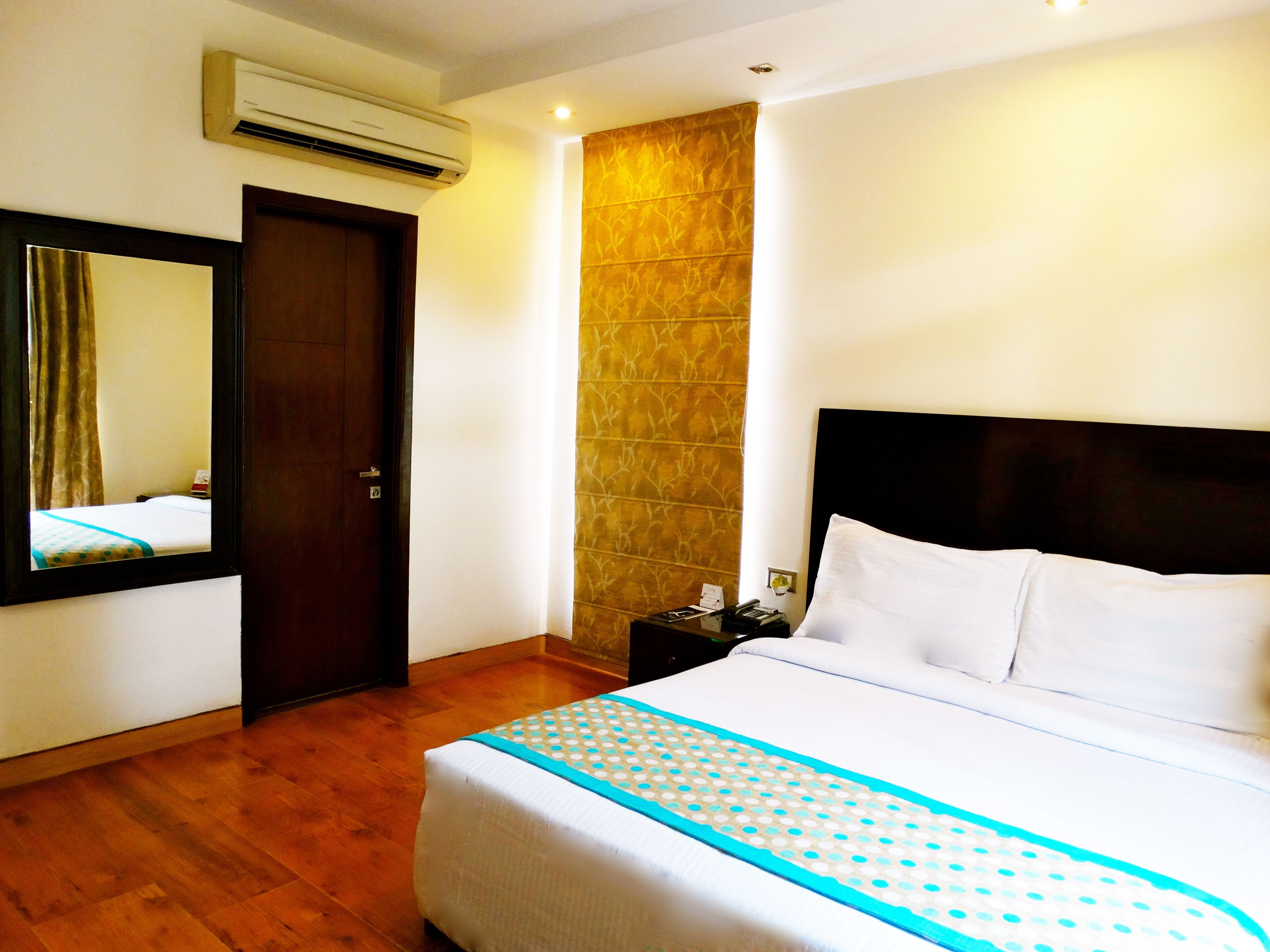 Habitación de hotel con cama, piso de madera, aire acondicionado instalado en la pared, una puerta y una mesita de noche con teléfono y lámpara.