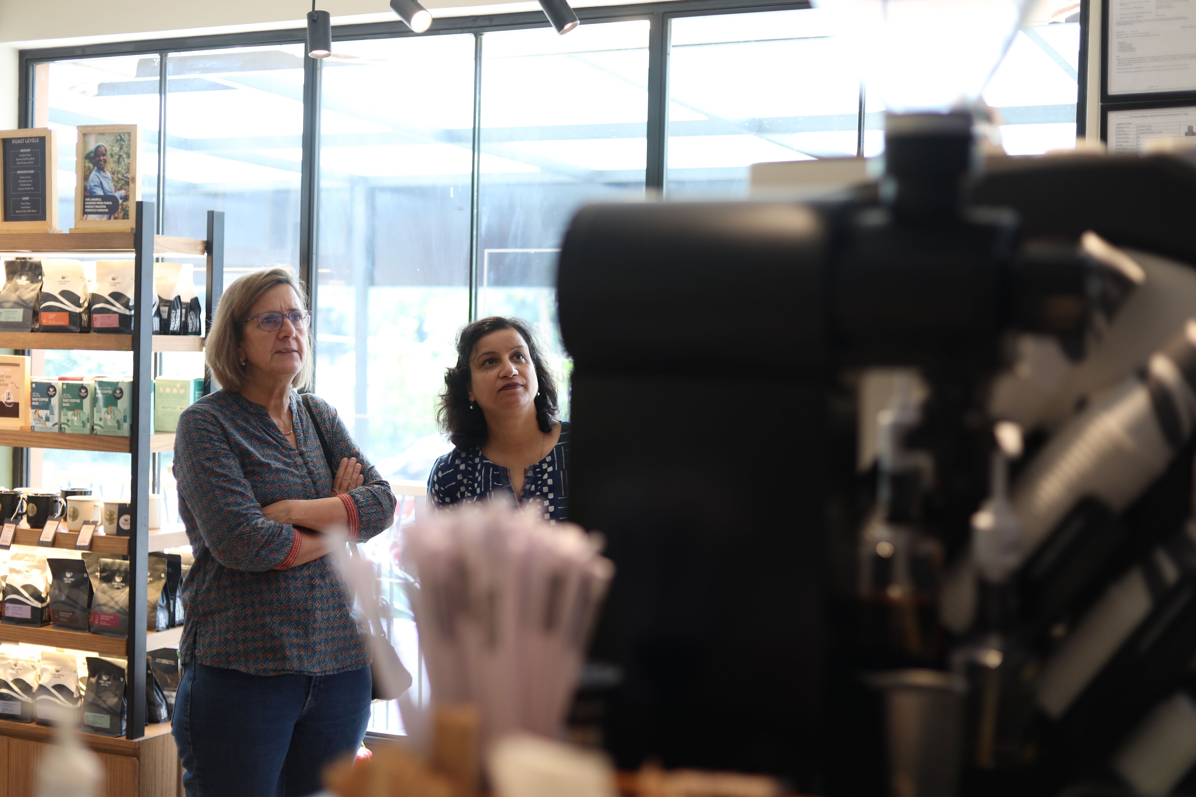Interior de una cafetería con una máquina de café, estantes con productos de café y dos personas de pie al frente.